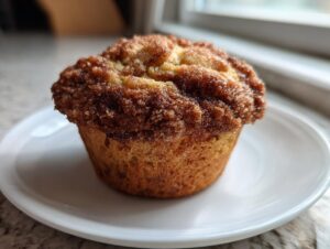 A close-up of a freshly baked sourdough discard cinnamon muffin with a thick, sugary cinnamon streusel topping, sitting on a small white plate.