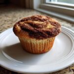 A freshly baked sourdough discard cinnamon muffin with a rich cinnamon swirl top, sitting on a white plate.