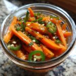 Close-up of bright orange pickled carrots sticks mixed with sliced green jalapeños and spices in a glass jar.
