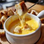 A piece of crusty bread is dipped into a white bowl of melted cheese fondue recipe, showing long, gooey cheese pulls.