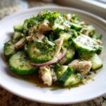 A close-up of a refreshing thai cucumber salad with chicken, featuring sliced cucumbers, herbs, and red onion in a savory dressing.