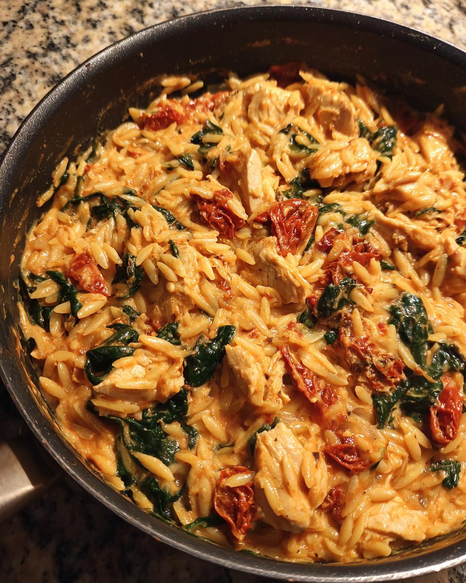 Close-up overhead view of creamy Tuscan chicken orzo with spinach and sun-dried tomatoes in a black skillet.