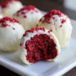 A close-up view of a white chocolate-dipped red velvet cake ball with a bite taken out, revealing the vibrant red interior.
