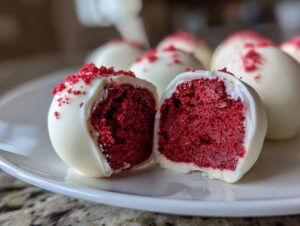 A white chocolate coated red velvet cake ball cut in half, revealing the rich red interior and red sprinkles.