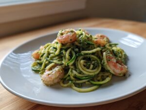 A close-up of zucchini noodles pesto shrimp tossed in vibrant green pesto sauce, served on a white plate.