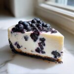 A perfect slice of creamy Blueberry Cheesecake with a dark cookie crust and fresh blueberry topping.
