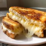 Close-up of a perfectly golden brown grilled cheese sandwich cut in half, showing dramatic, gooey, melted cheese stretching between the slices.