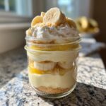 A layered banana pudding parfait in a mason jar topped with whipped cream and fresh banana slices.