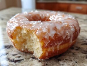 A close-up photo showing a bite taken out of a fluffy, golden Glazed yeast donut sitting on a speckled countertop.