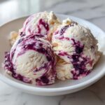 Three scoops of creamy vanilla Blueberry swirl ice cream with rich purple berry ribbons served in a white bowl.