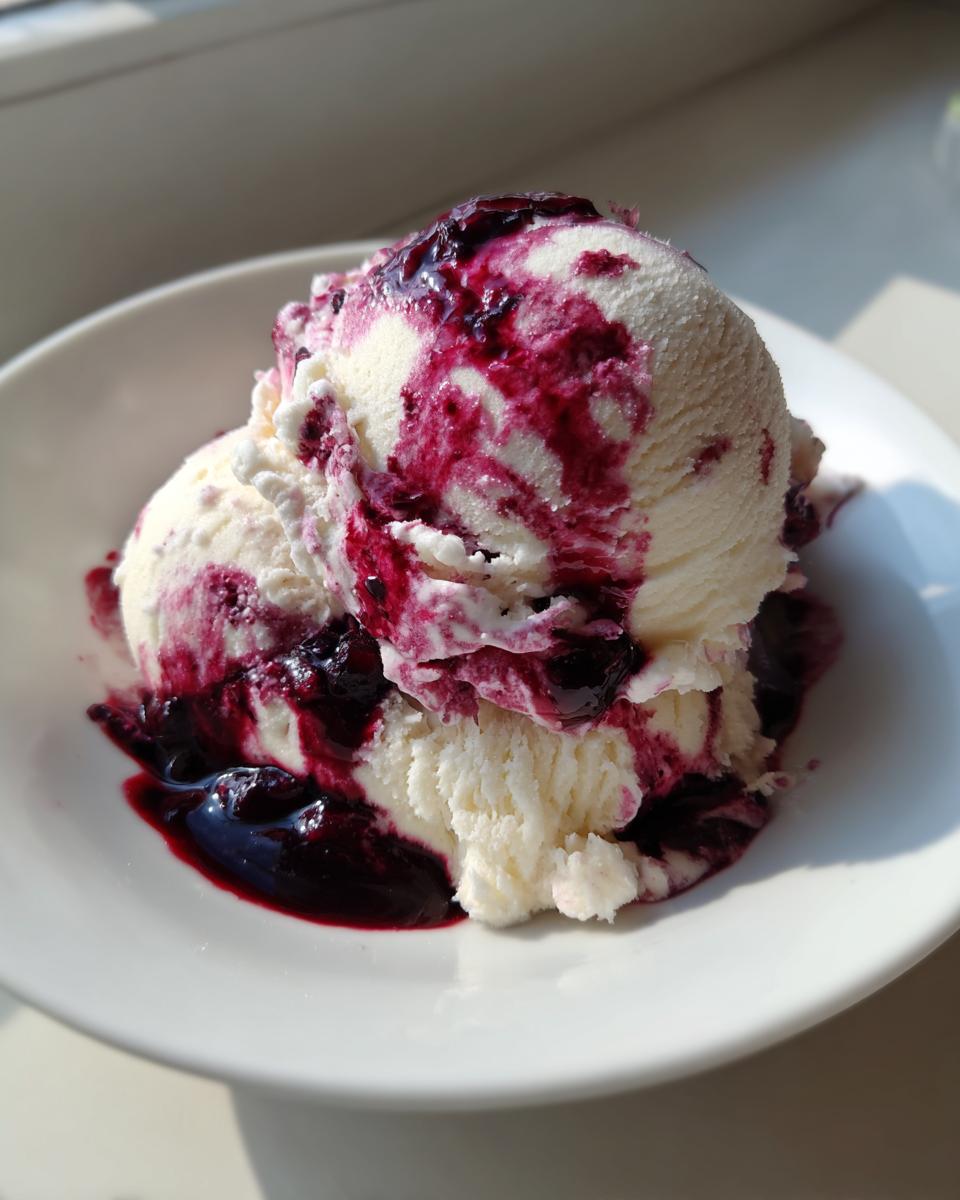 Two scoops of vanilla ice cream generously topped with a dark purple blueberry swirl sauce in a white bowl.