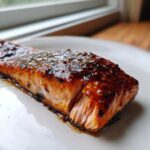 Close-up of a perfectly cooked fillet of Bourbon glazed salmon with a thick, shiny, dark glaze on a white plate.
