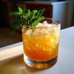 A vibrant orange Bourbon peach smash cocktail served over crushed ice and garnished with fresh mint.