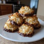 A stack of golden brown coconut macaroon cookies dipped in dark chocolate resting on a white plate.
