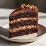 A tall slice of decadent chocolate hazelnut cake with layers of dark chocolate sponge and hazelnut filling, topped with frosting and chopped nuts.