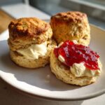 Three freshly baked clotted cream scones served on a plate, one split and topped with clotted cream and strawberry jam.