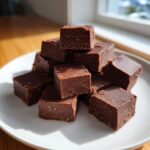A pile of rich, square pieces of dark chocolate fudge stacked on a white plate near a window.