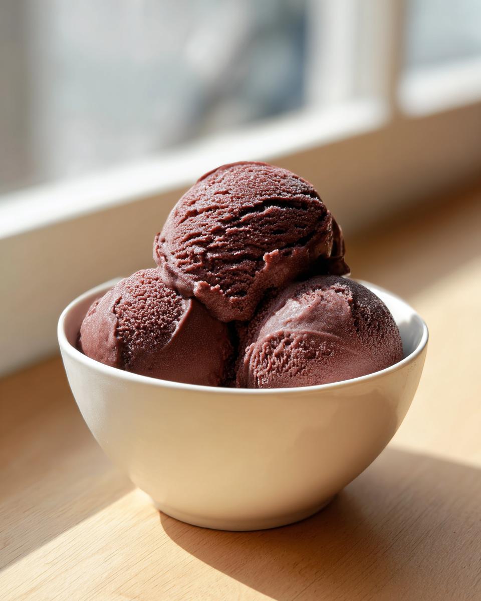 Three scoops of rich, dark chocolate sorbet piled high in a white bowl, illuminated by sunlight.