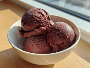 Three scoops of intensely colored dark chocolate sorbet served in a small white bowl, illuminated by sunlight.