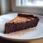 A dense, rich slice of Flourless chocolate torte dusted generously with cocoa powder, served on a white plate.