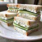 Four crustless cucumber sandwiches stacked on a white plate, featuring thick cucumber slices and herbed cream cheese filling.