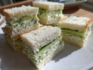 Close-up of several crustless cucumber sandwiches stacked on a white plate, showing creamy filling and fresh cucumber slices.