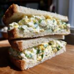 Two halves of a thick egg salad sandwich stacked on a wooden board, showing creamy filling with green herbs.