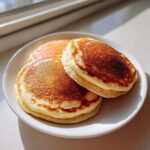 Three golden-brown, fluffy Applesauce pancake stacked slightly on a white plate, illuminated by bright sunlight.