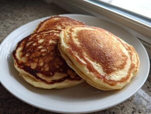 Three golden brown, fluffy Applesauce pancake stacked slightly on a white plate near a window.