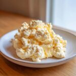 A close-up of fluffy, pale yellow cottage cheese scrambled eggs piled high on a speckled white plate.