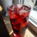 Close-up of a tall, sweating glass filled with deep red Hibiscus iced tea and large ice cubes.