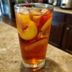 A tall, sweating glass filled with cold Peach iced tea, ice cubes, and slices of fresh peach.