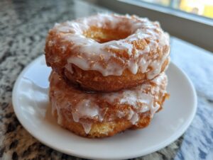 Two perfectly textured, glazed old fashioned cake donuts stacked on a small white plate near a window.