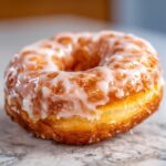 A close-up, appetizing shot of a single, golden-brown glazed yeast donut resting on a marble surface.