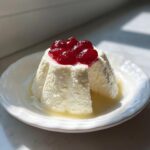 A molded serving of creamy Greek yogurt panna cotta topped with bright red berry compote, sitting in a pool of liquid on a white plate.