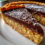 Close-up of a slice of Hazelnut praline tart showing a thick, textured filling, topped with dark chocolate ganache and flaked coconut.