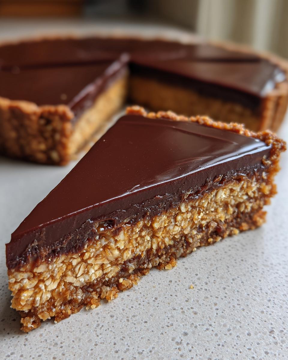 A close-up slice of Hazelnut praline tart showing a crunchy oat base, praline filling, and shiny chocolate topping.