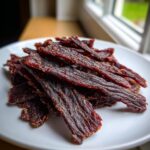 Close-up of a pile of dark, seasoned strips of homemade beef jerky resting on a white plate.