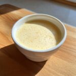 A white bowl filled with creamy, pale yellow Homemade vanilla pudding, showing small bubbles on the surface, set on a wooden board.