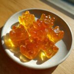 A small plate holding several translucent, amber-colored honey gummy bear candies glistening in the sunlight.
