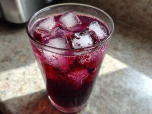 Close-up of a tall glass filled with vibrant, deep purple Blackberry sage tea over ice cubes, showing condensation.
