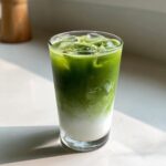 A tall glass showing distinct layers of white milk and bright green matcha milk tea with ice cubes.