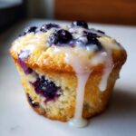 A close-up of a moist Lemon blueberry cupcake topped with fresh blueberries and a thick, dripping lemon glaze.