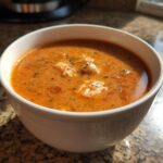 Close-up of a steaming white bowl filled with rich, orange-red lobster bisque, topped with herbs and chunks of lobster meat.