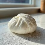 A smooth, white ball of freshly made marshmallow fondant dusted with powdered sugar on a marble surface.