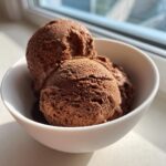 Two scoops of dark, rich Mexican chocolate ice cream served in a white bowl, illuminated by sunlight.