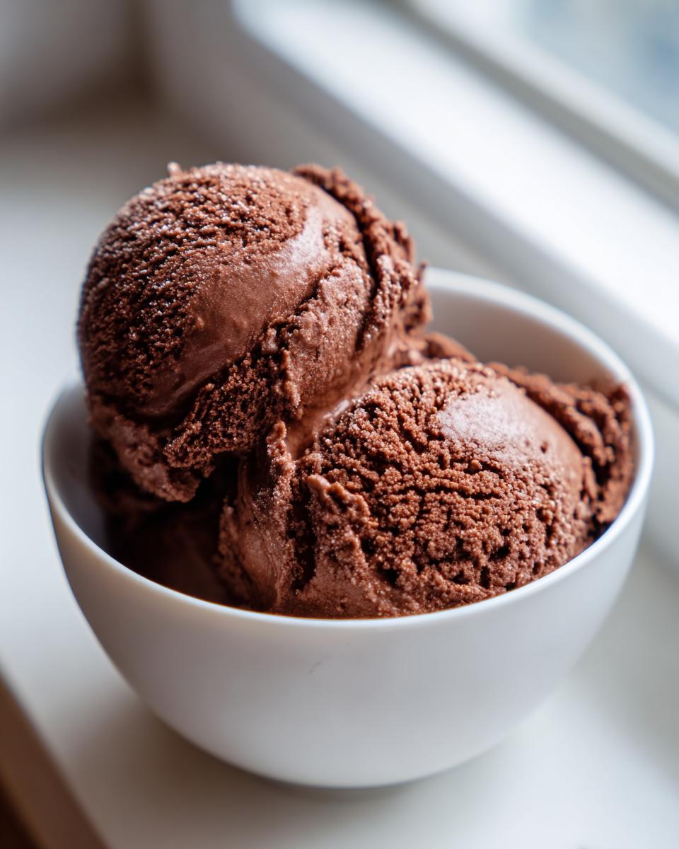 Three scoops of rich, dark brown Mexican chocolate ice cream served in a white bowl near a bright window.