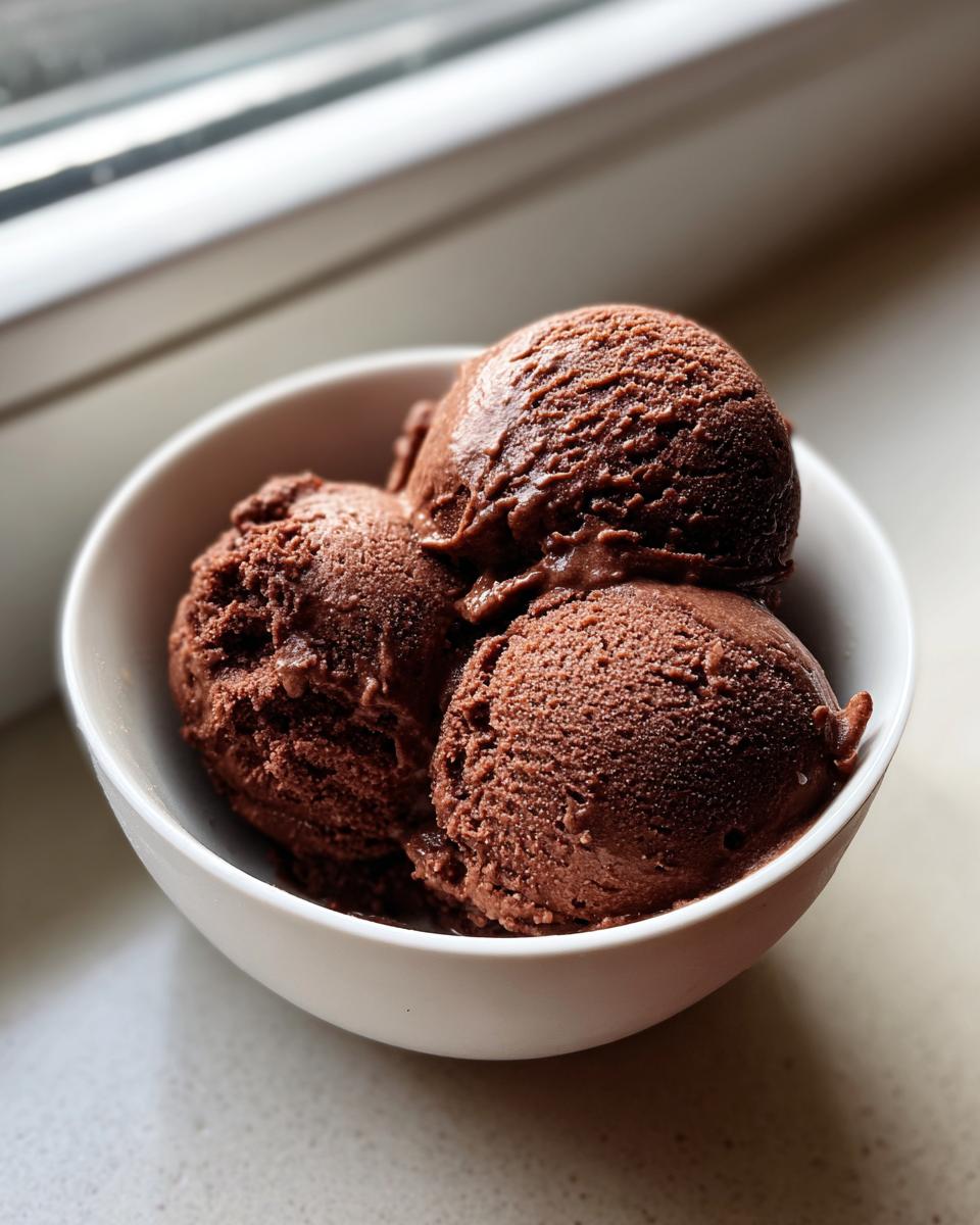Three scoops of rich, dark brown Mexican chocolate ice cream served in a white bowl near a window.