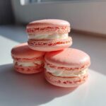 Three perfectly piped pink strawberry macaroon cookies filled with white cream stacked on a white surface.