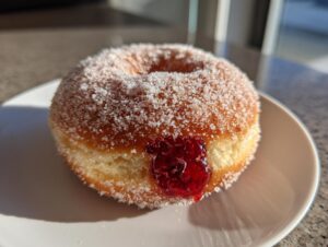 A close-up of a freshly made raspberry jelly donut, coated in sugar, with bright red filling visible.